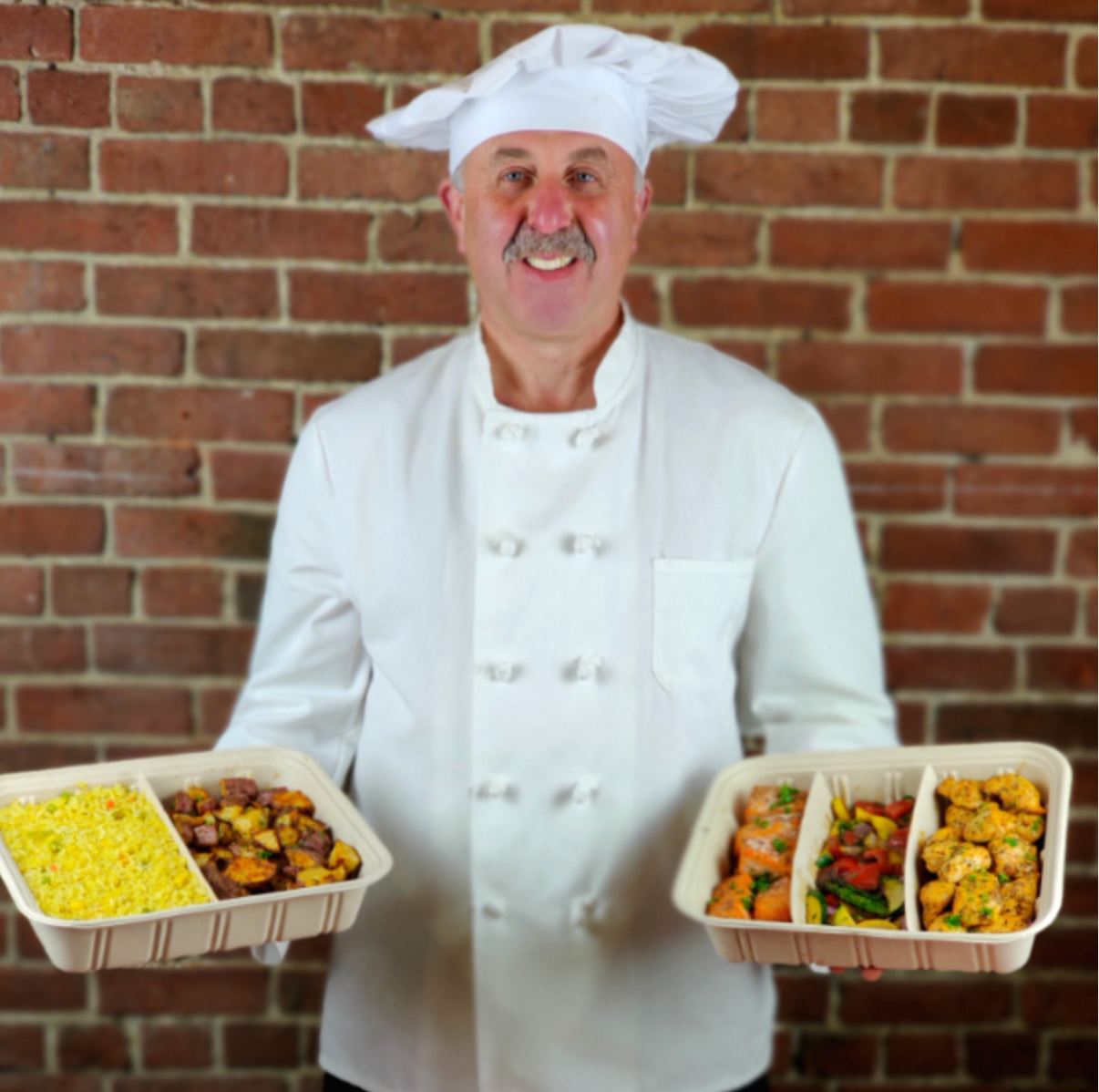a chef holding two trays of food in front of a brick wall.