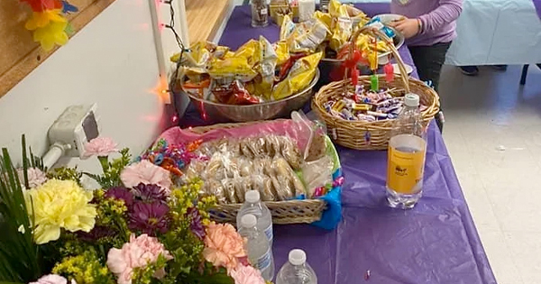 A table draped in a purple tablecloth showcases an inviting spread of snack bags, sandwiches, wrapped candies, and water bottles. A bouquet of fresh flowers adds a charming touch to the display. In the background, someone is tidying up the arrangement, making sure everything looks perfect.