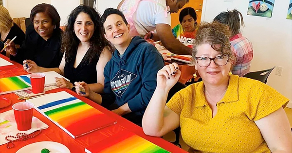 A cheerful group sits around a table full of art supplies, each person with a paintbrush in hand, joyfully painting and sharing smiles.