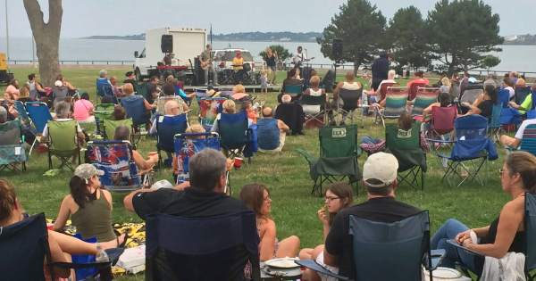 A lively crowd relaxes on lawn chairs and blankets, enjoying an outdoor concert in the park by the water, even under the cloudy sky.