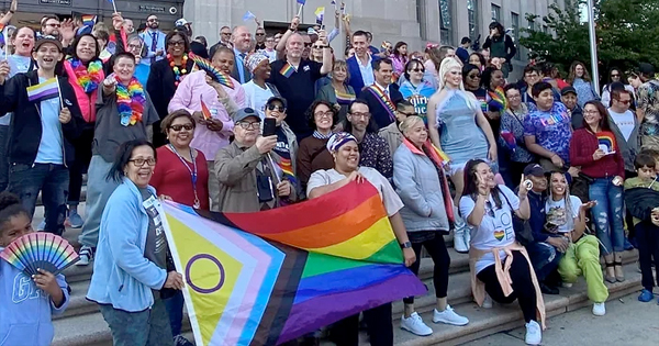 A vibrant group of individuals gathers on steps, proudly displaying rainbow flags and donning colorful outfits, celebrating an exciting outdoor event.