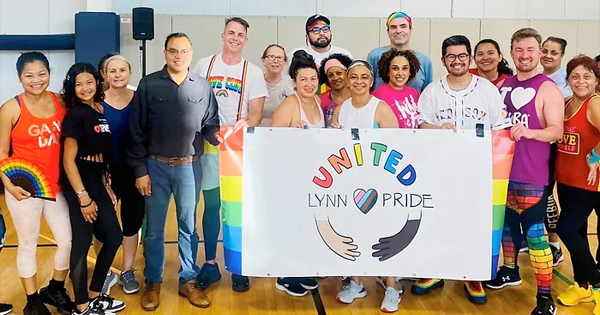 In a vibrant gym setting, an inclusive group proudly holds a "United Lynn Pride" banner adorned with rainbow designs. The participants, dressed in colorful attire and accessories, celebrate unity and diversity.