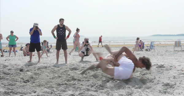 Two individuals are engaged in a playful wrestling match on a sandy beach, drawing the attention of bystanders who snap photos. In the background, sunbathers relax on their beach chairs, adding to the lively seaside scene.