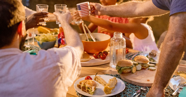 A cheerful group cheers with their drinks around an outdoor table brimming with delicious food like burgers, corn on the cob, fresh salad, and a pitcher of cool beverages.