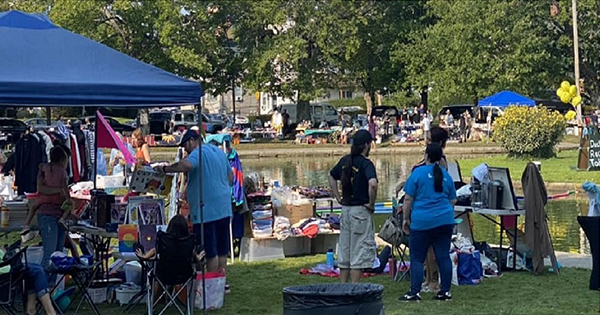 People shopping and selling goods at an outdoor market set up with tables and tents on a grassy area by the pond.