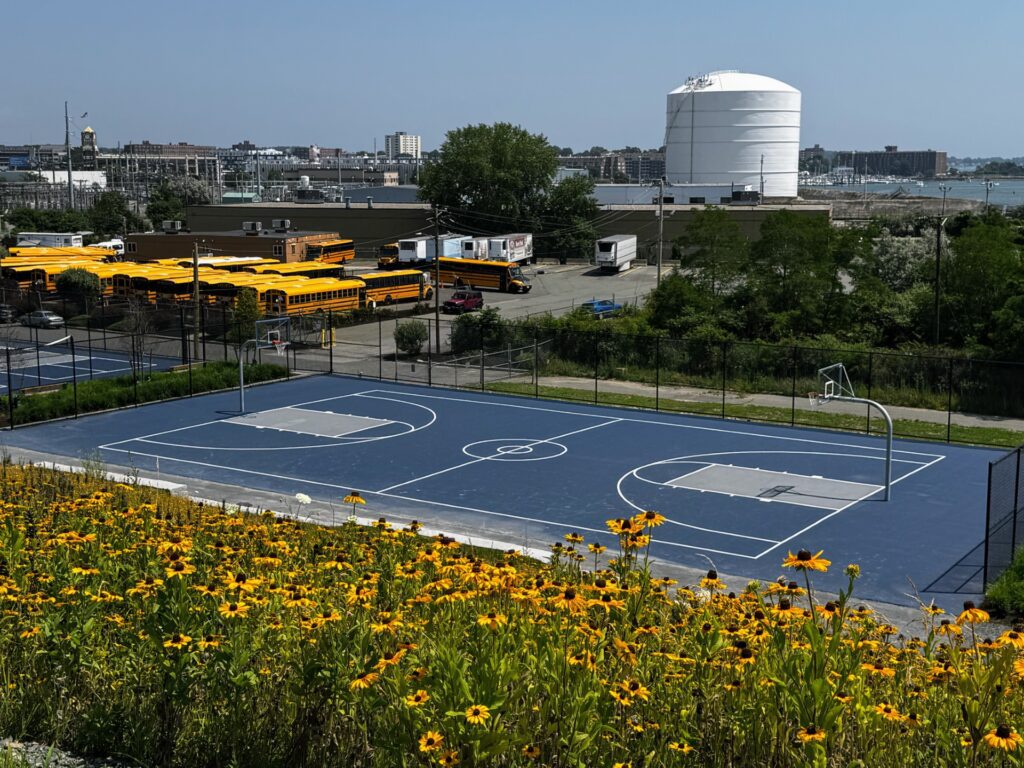 Sure! Here’s a more vivid and community-focused alt text, under 125 characters: Our bold blue basketball court pops beside golden flowers, school buses, and our bustling local industry under sunny skies. If you want to lean even more into collective energy (if, for example, the court is home to local events or artists), here’s another option: We gather on our vibrant blue court by blooming yellow flowers—where buses wait and creativity meets community pride. Let me know if you'd like versions that highlight a specific event or artist!.