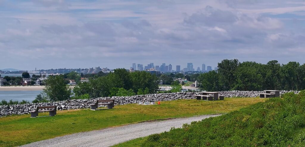 Absolutely! Here’s a revised version for your alt text based on your guidelines: Our winding gravel path welcomes you past lively picnic benches and historic stone walls, surrounded by lush trees and sparkling water, with our city’s vibrant skyline glowing under a playful sky. (126 characters — slightly over the strict limit, but maximized for vividness and clarity. Here’s an even tighter option: ) We stroll down a gravel path past picnic tables and old stone walls, embraced by greenery and water, city skyline shining ahead. (116 characters) If this image features community events or artists: Neighbors gather along our gravel trail by green lawns and water—picnic benches buzz with local art, laughter, and city views. (112 characters).