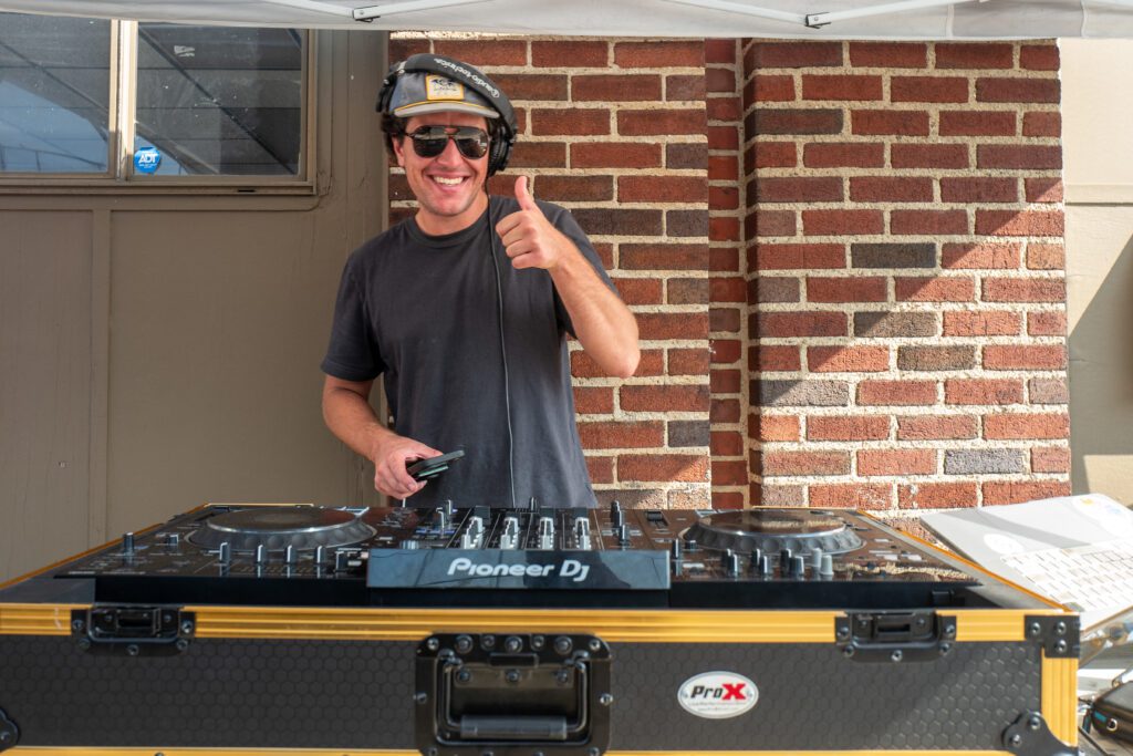 A lively DJ in sunglasses and headphones beams, giving a thumbs-up behind Pioneer DJ gear, energizing the Essex County scene.
