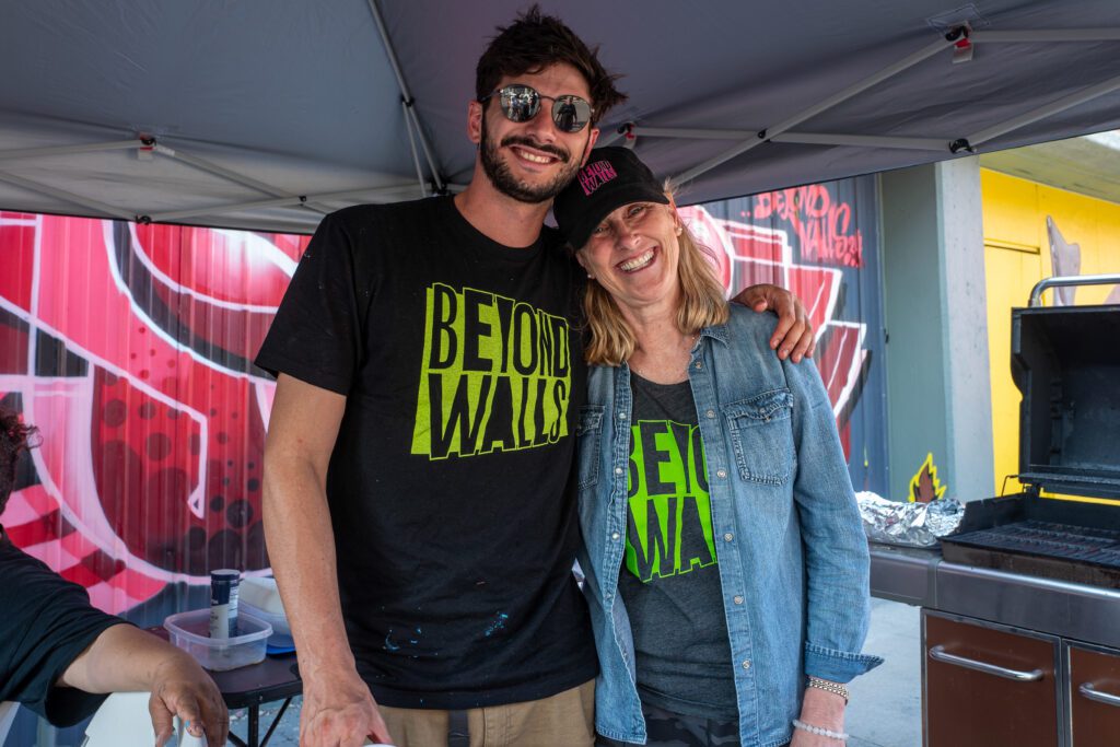 Smiling in "Beyond Walls" shirts, two people unite under a canopy near sizzling grills and foil trays, with a lively mural celebrating Essex County’s creative community behind them.