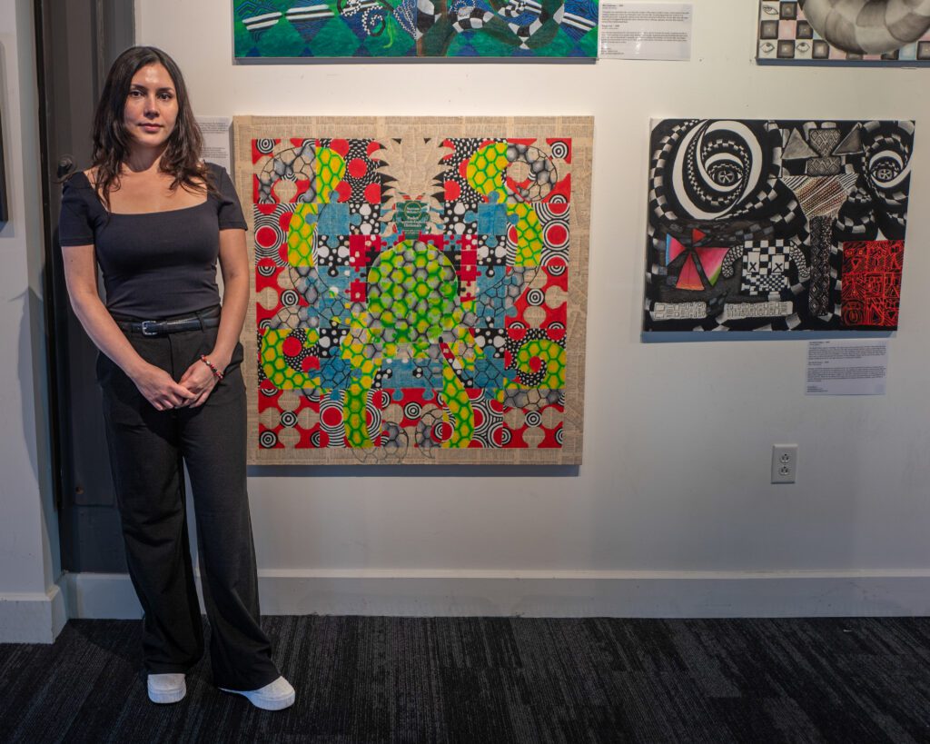 A woman beams indoors beside a lively abstract painting bursting with bold colors, surrounded by local art that sparks Essex County pride.