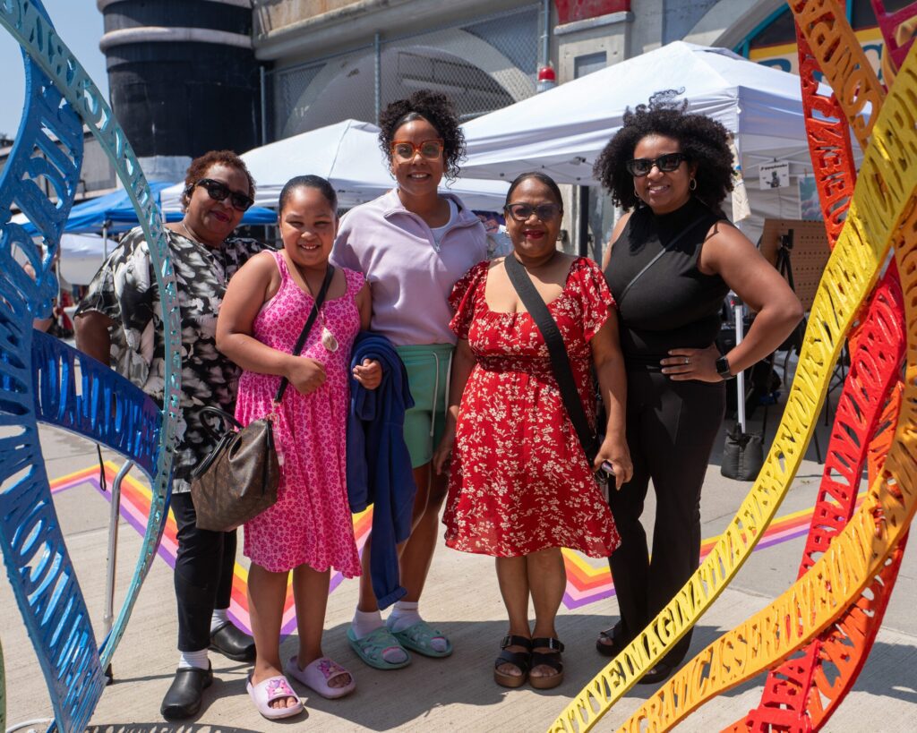 Five smiling people gather at Lynn Arts Festival 2025, basking in colorful Essex County art, lively tents, and a joyful community vibe.