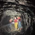 Three adventurers explore Lynn's Dungeon Rock, flashlight in hand, their teamwork lighting the rugged stone walls around them.