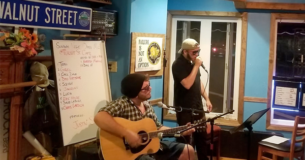 Local musicians energize an indoor crowd; one strums guitar, another sings. Setlist and Walnut Street sign nod to Essex County spirit.