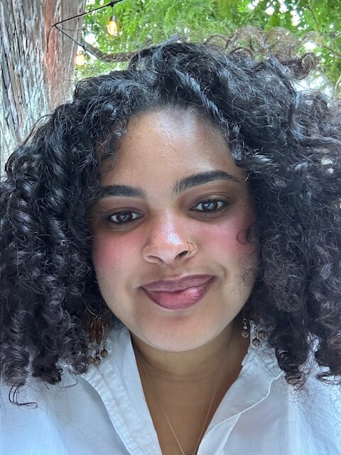 A vibrant individual with curly dark hair and a nose ring beams outdoors in Essex County, surrounded by lush greenery and festive lights.
