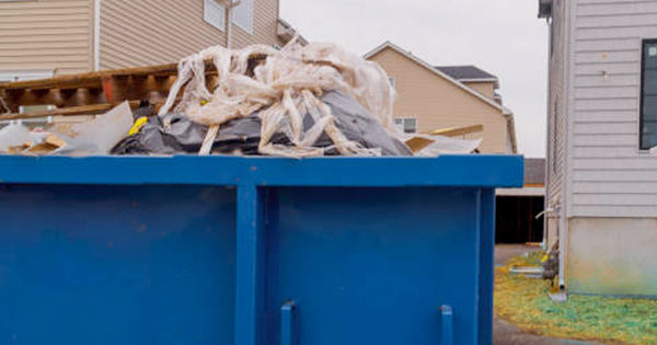 A blue dumpster filled with construction debris and plastic sheeting sits between two residential houses on a cloudy day.