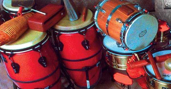 A group of red conga drums, a snare drum, and various percussion instruments including a guiro, cowbell, and maracas are arranged together on a floor.