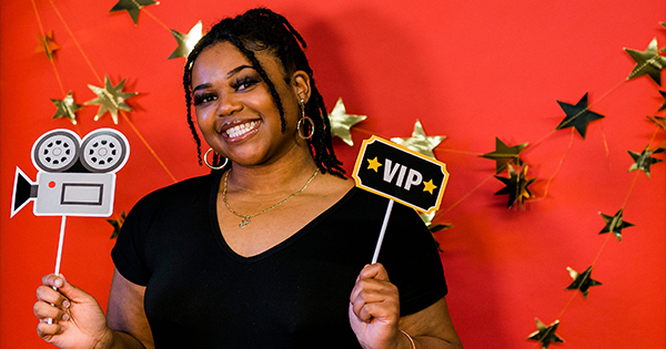 A woman stands in front of a red background decorated with gold stars, holding a paper cutout of a vintage film camera in one hand and a sign reading "VIP" in the other. She is smiling and wearing a black shirt.