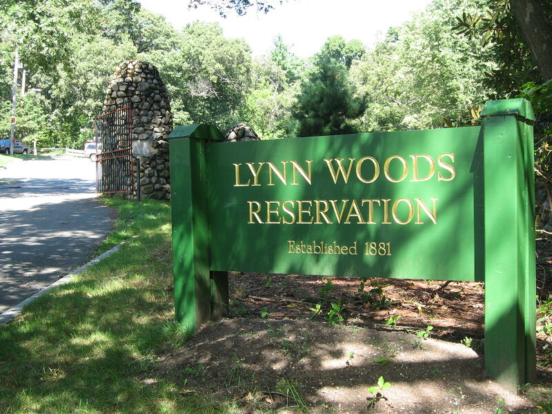 A green wooden sign reading "Lynn Woods Reservation, Established 1881" stands near a paved road with a stone gate entrance and dense trees in the background.