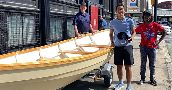 Three people stand on a city sidewalk next to a beige rowboat on a trailer. One holds a cap, another wears a red shirt and patterned pants, and the third stands behind the boat. A building and street signs are visible in the background.