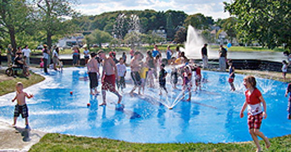 Children and adults play in a splash pad water park on a sunny day. Water sprays from jets in the blue ground. Trees, a fountain, and a pond are visible in the background. People are dressed in summer clothes and swimsuits.