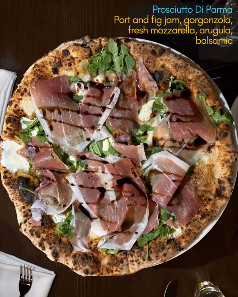 A wood-fired pizza topped with prosciutto, arugula, gorgonzola, fig jam, mozzarella, and a drizzle of balsamic. The pizza is on a dark table with white napkins and cutlery visible at the edges. Text with the pizza’s ingredients appears in the corner.