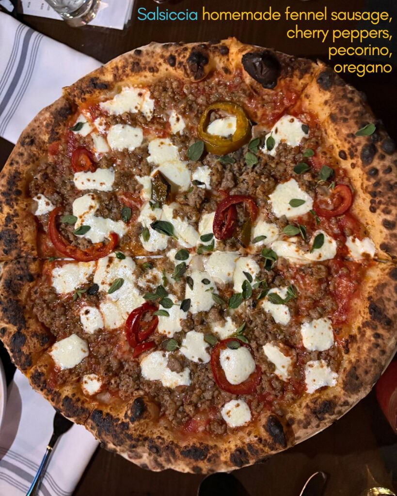 A wood-fired pizza topped with pieces of sausage, sliced cherry tomatoes, melted cheese, yellow and red peppers, and fresh oregano, served on a wooden table with a napkin and a drink visible nearby.