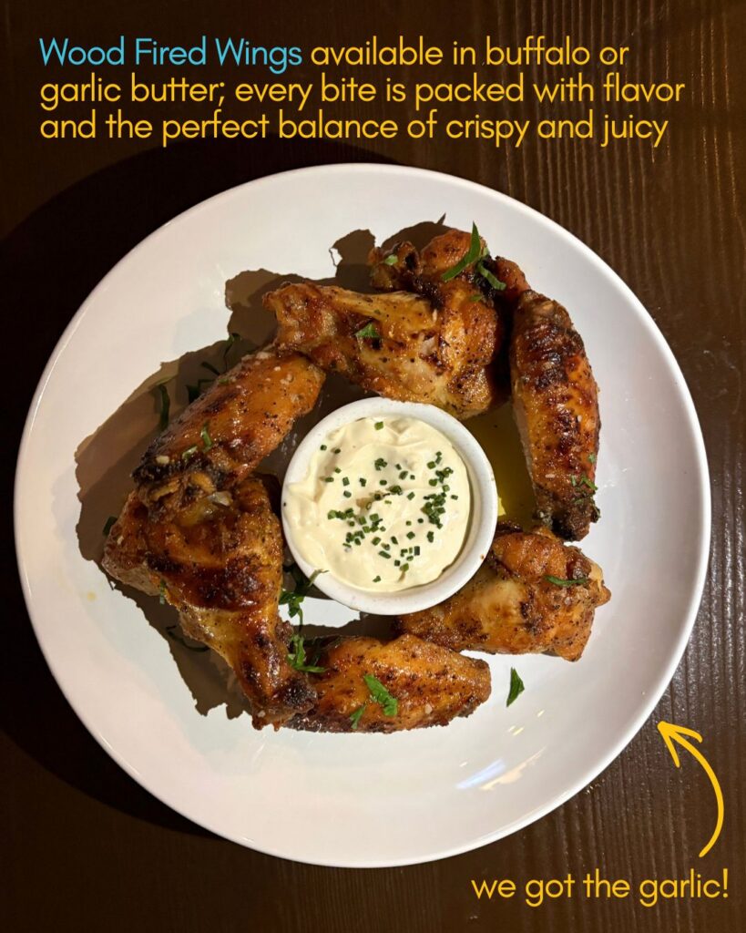 A white plate with six wood-fired chicken wings arranged around a cup of creamy dipping sauce, garnished with herbs, sits on a brown table. Text on the image describes the wings as available in buffalo or garlic butter.