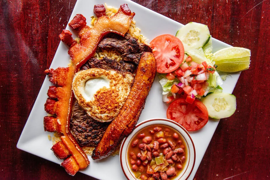 A plate with steak, fried egg, bacon, sausage, fried plantain, rice, lettuce, tomato, cucumber, lime wedges, pico de gallo, and a small bowl of beans at Casa Antigua in Downtown Lynn MA