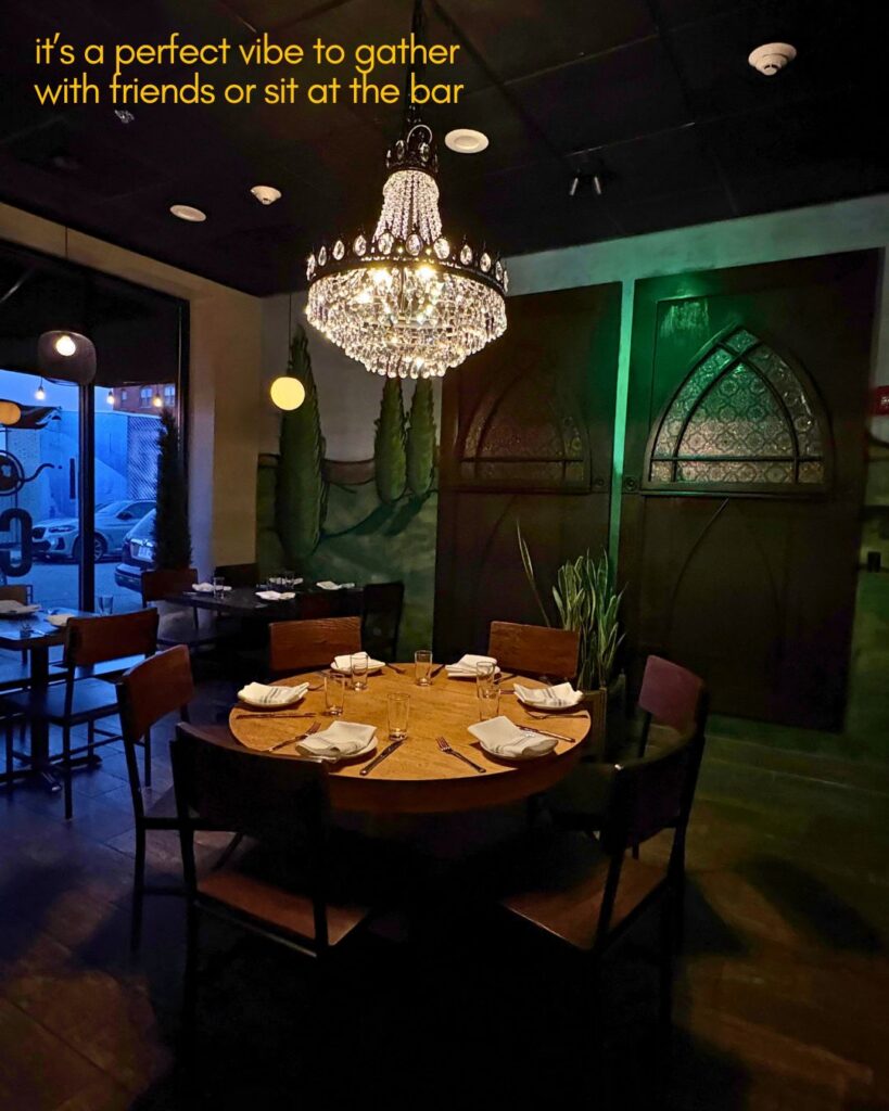 A round dining table set for six with napkins and glasses sits under a large chandelier in a dimly lit restaurant. Dark wood chairs surround the table. Green plants, stained glass doors, and large windows are visible in the background.