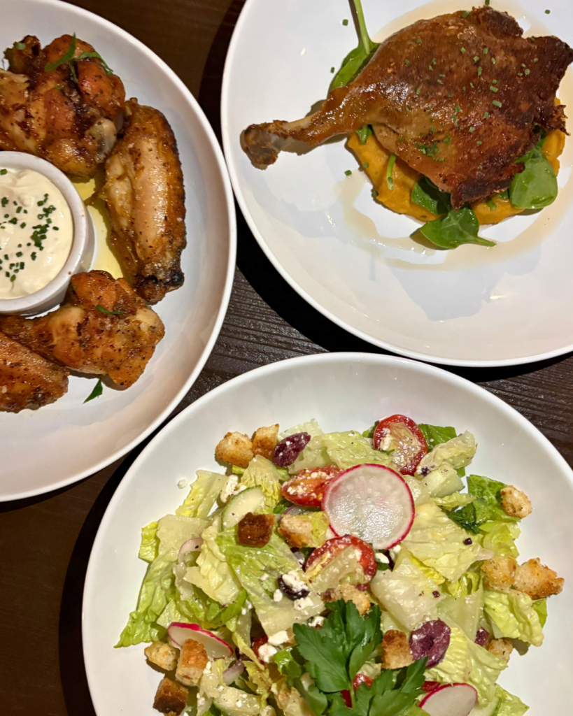 Three white plates on a dark table: one with chicken wings and dipping sauce, one with crispy duck leg on mashed vegetables, and one with a green salad topped with radishes, olives, croutons, and cheese.