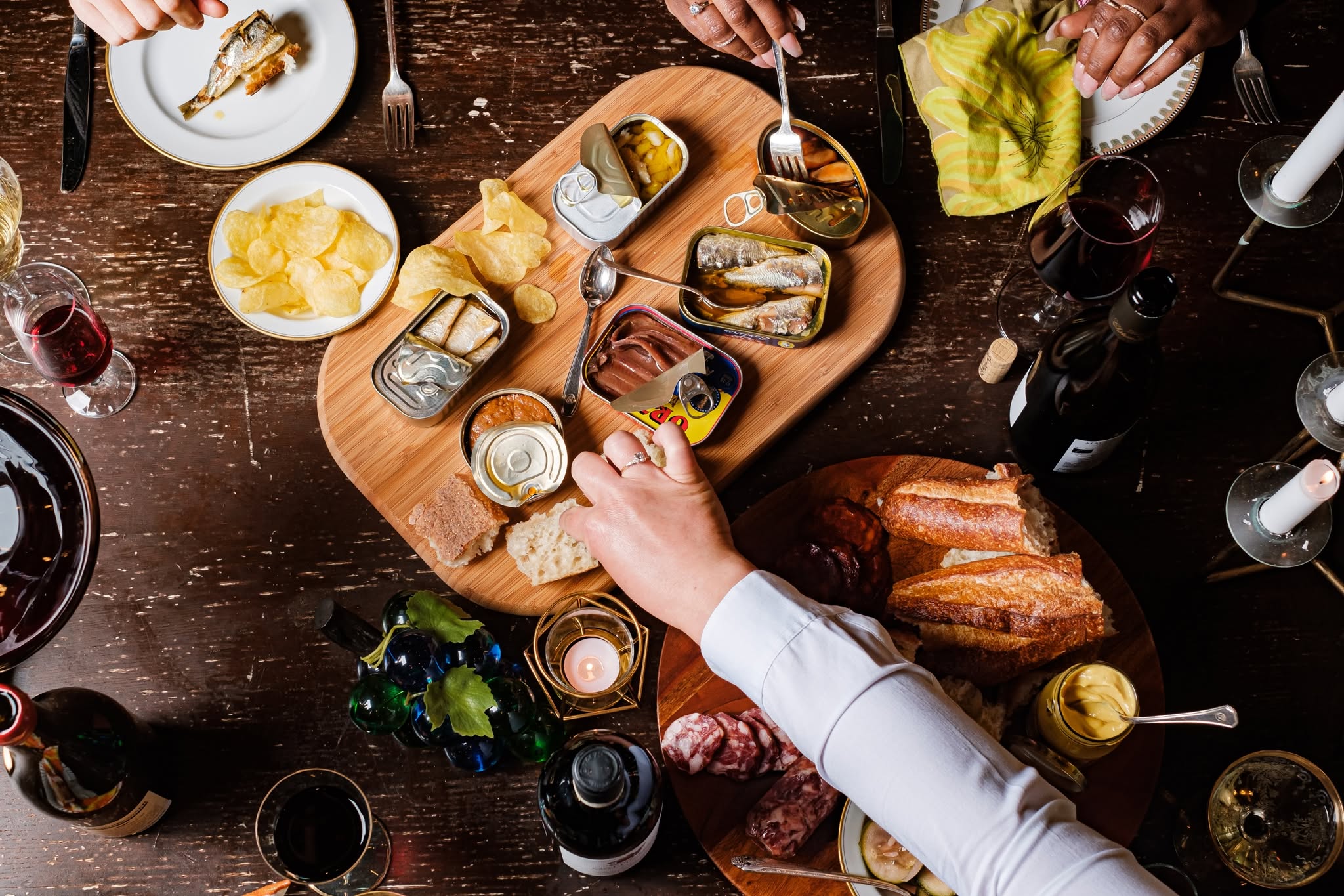 A dinner table with an assortment of tinned fish, bread, crackers, chips, cured meats, condiments, wine, and candles. Several people’s hands reach for food, and plates, glasses, and utensils are visible around the wooden boards at Lucille's in Downtown Lynn, MA.