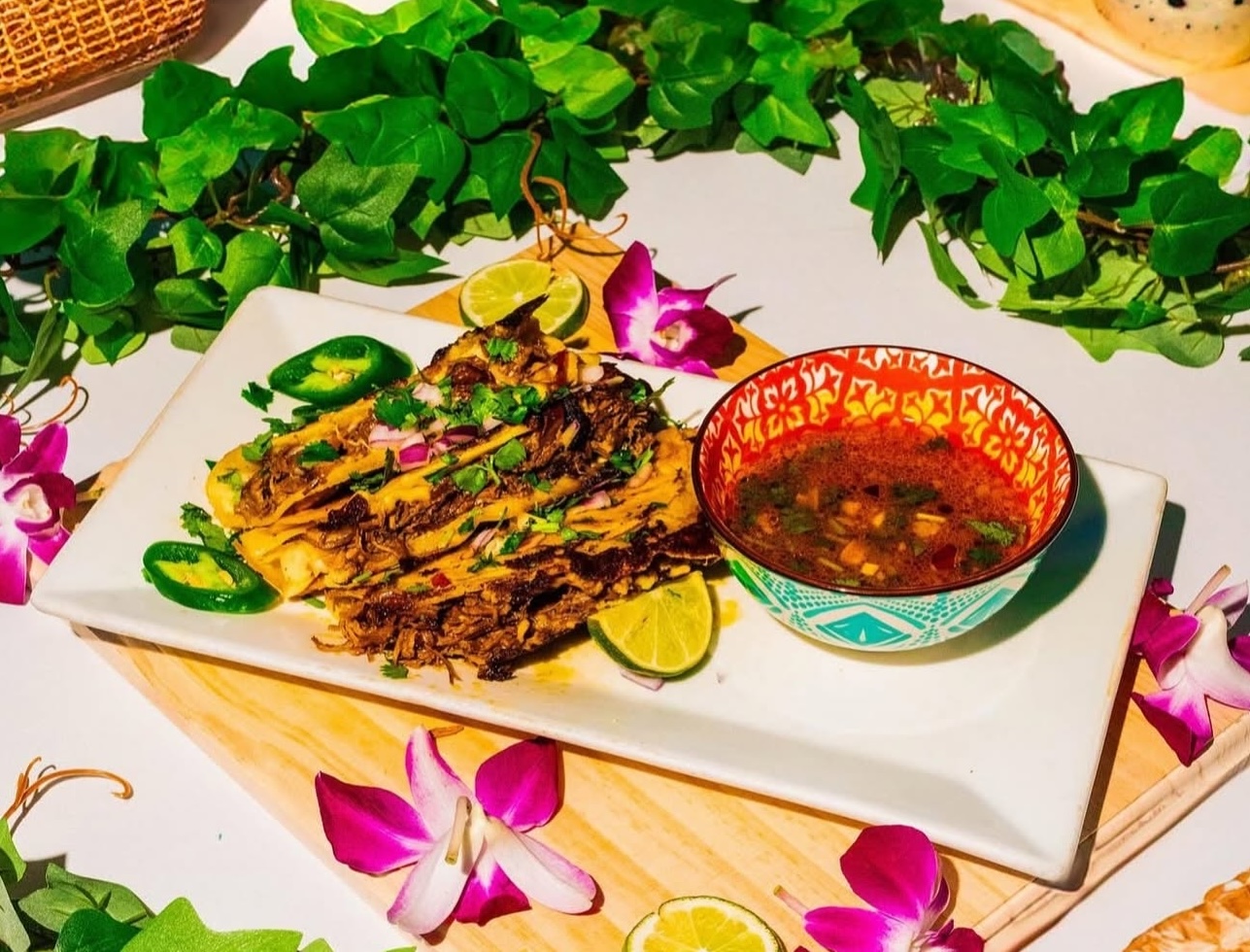A rectangular plate holds birria tacos with lime wedges, jalapeño slices, and cilantro, next to a small bowl of birria at Estefani's in Downtown Lynn MA