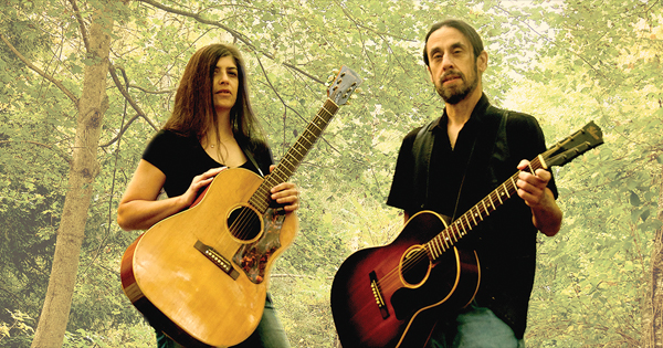 A man and a woman holding acoustic guitars stand in a wooded area.